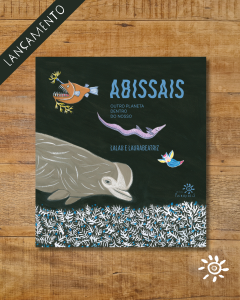 Capa do livro *Abissais – Outro Planeta Dentro do Nosso*, de Lalau e Laurabeatriz, publicado pela Editora Peirópolis. A capa tem fundo escuro e apresenta uma ilustração colorida de animais marinhos das profundezas do oceano, incluindo um peixe-pescador de dentes afiados, um peixe longo e rosado, uma baleia em tons de cinza e pequenos peixes brilhantes. Na parte inferior, há uma representação de corais brancos com detalhes azuis. O título *Abissais* está em azul, e o subtítulo "Outro planeta dentro do nosso" aparece em branco logo abaixo. A capa está posicionada sobre um fundo de madeira. No canto superior esquerdo da imagem, há uma faixa preta diagonal com a palavra "LANÇAMENTO" em branco, e no canto inferior direito, um ícone de sol estilizado, ambos sobre o fundo de madeira.