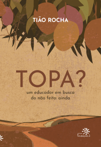 Capa do livro Topa? Um educador em busca do não feito: ainda, de Tião Rocha, publicado pela Editora Peirópolis. O título aparece em letras grandes no centro da imagem, com o subtítulo logo abaixo, em menor tamanho. O fundo tem textura terrosa e tons quentes, com ilustração de um caminho sinuoso atravessando uma paisagem de colinas. Na parte superior, há ramos de uma mangueira com frutas maduras em tons de laranja, amarelo e roxo, contrastando com o fundo bege. Nos cantos inferiores esquerdo e direito está o logotipo da Editora Peirópolis.