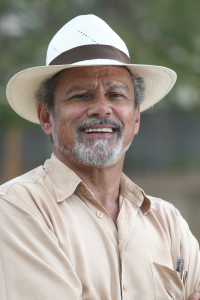 Foto de Tião Rocha, educador e antropólogo cultural. Ele está sorrindo, vestindo uma camisa bege de mangas compridas e um chapéu de palha com faixa marrom escura. Tem barba e cabelo grisalhos. No bolso da camisa, há algumas canetas. O fundo é desfocado, com tons de verde e cinza, sugerindo um ambiente ao ar livre.
