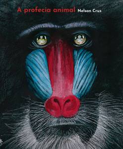 Capa do livro A profecia animal, de Nelson Cruz. A imagem mostra em close o rosto de um mandril, com destaque para suas cores vivas: o nariz vermelho vibrante e as laterais do rosto em azul. O fundo é escuro, o que realça ainda mais os traços faciais do animal. Nos olhos do mandril, vê-se o reflexo do autor, desenhando. No canto superior esquerdo, em letras vermelhas e brancas, está o título do livro e o nome do autor: “A profecia animal — Nelson Cruz