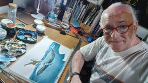 O autor e ilustrador Nelson Cruz sorri levemente para a câmera em seu ateliê. Ele usa óculos de armação vermelha e uma camiseta clara com estampa. Está sentado diante de uma mesa de trabalho repleta de tintas, pincéis, potes e paletas de pintura. À sua frente, sobre a mesa, há uma ilustração em aquarela em andamento, que mostra uma girafa equilibrando-se sobre uma baleia em meio ao oceano. Ao fundo, há uma estante cheia de livros, materiais de arte e objetos de trabalho, compondo um ambiente criativo e acolhedor.